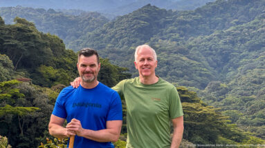 Two trekkers posing shoulder to shoulder with a walking stick before a steep, densely forested ridge.