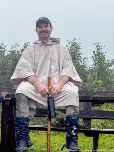 Seated trekker wearing a rain poncho and gaiters, holding a wooden walking stick against a wet green backdrop.