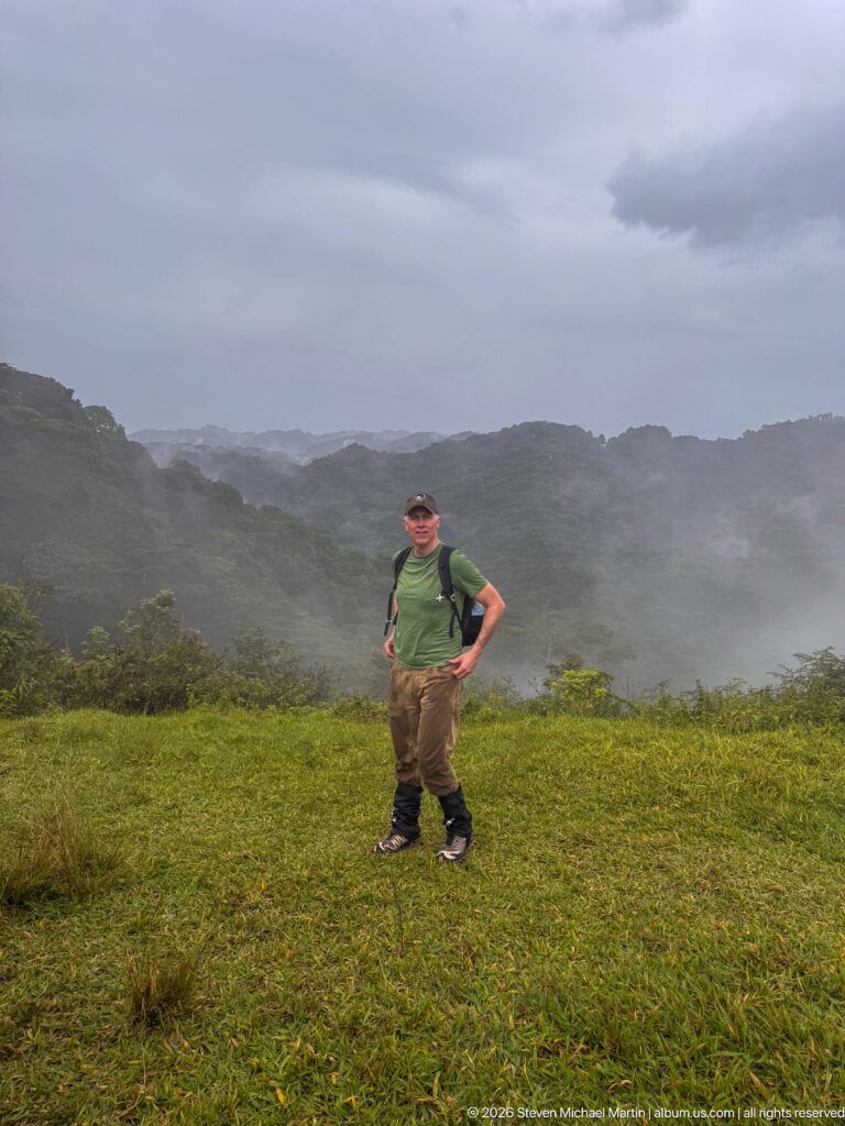 Trekker standing on a grassy ridge as rain and mist obscure the layered mountain landscape behind.