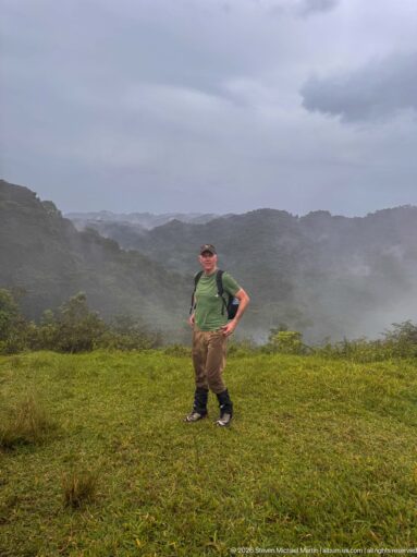 Trekker standing on a grassy ridge as rain and mist obscure the layered mountain landscape behind.