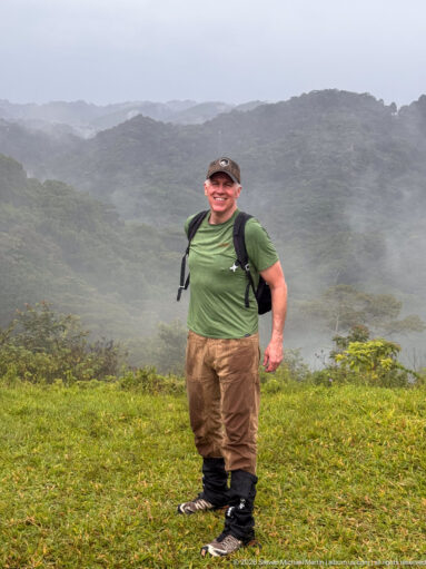 Smiling trekker with backpack and gaiters stands on a grassy clearing before mist drifting across forested hills.