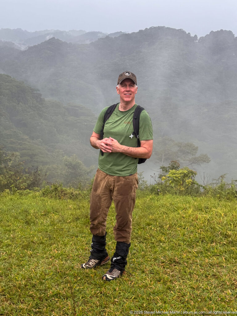 Man wearing hiking gear and backpack stands on grass before misty, densely forested mountain ridges.
