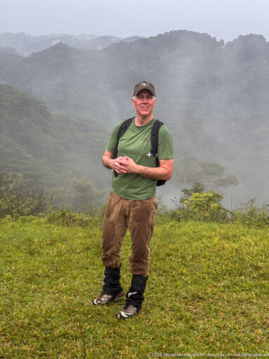 Man wearing hiking gear and backpack stands on grass before misty, densely forested mountain ridges.