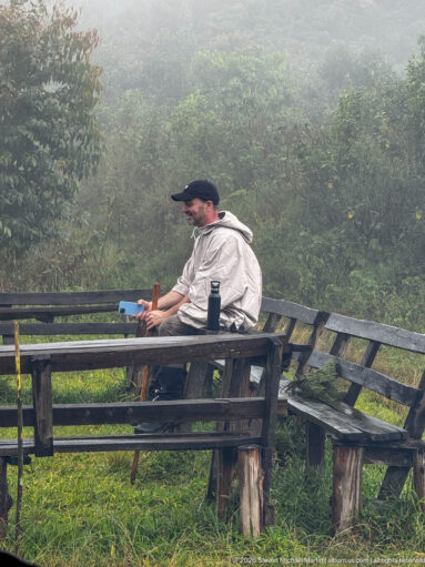 Man in a light rain poncho sits on a wooden bench beside trekking gear in dense misty forest.