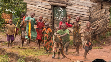 Smiling children wave and walk along a red-earth path beside a weathered wooden building in a green rural setting.