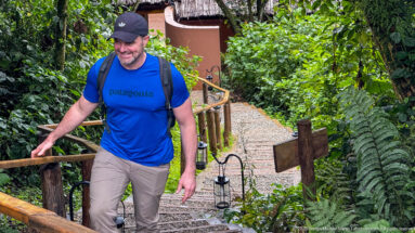Man with backpack and trekking pole climbs stone steps bordered by wooden rails and dense forest vegetation.