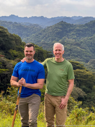 Two visitors posing with a walking stick on an open slope above deeply forested mountain ridges.