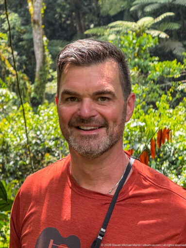 Smiling man in a red shirt standing in front of softly blurred green forest vegetation.