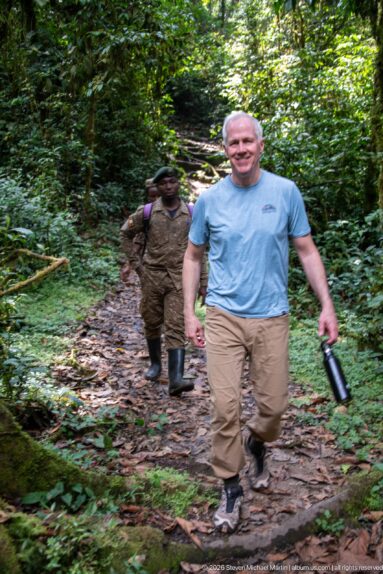 Hiker in blue shirt walking uphill on a muddy forest path with a uniformed guide behind him.