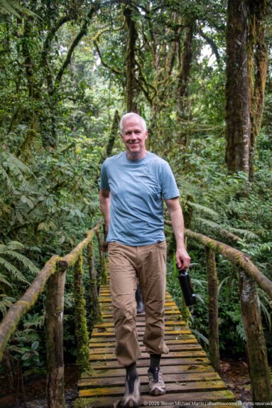 Man standing on a narrow wooden footbridge with rustic rails in dense green forest.