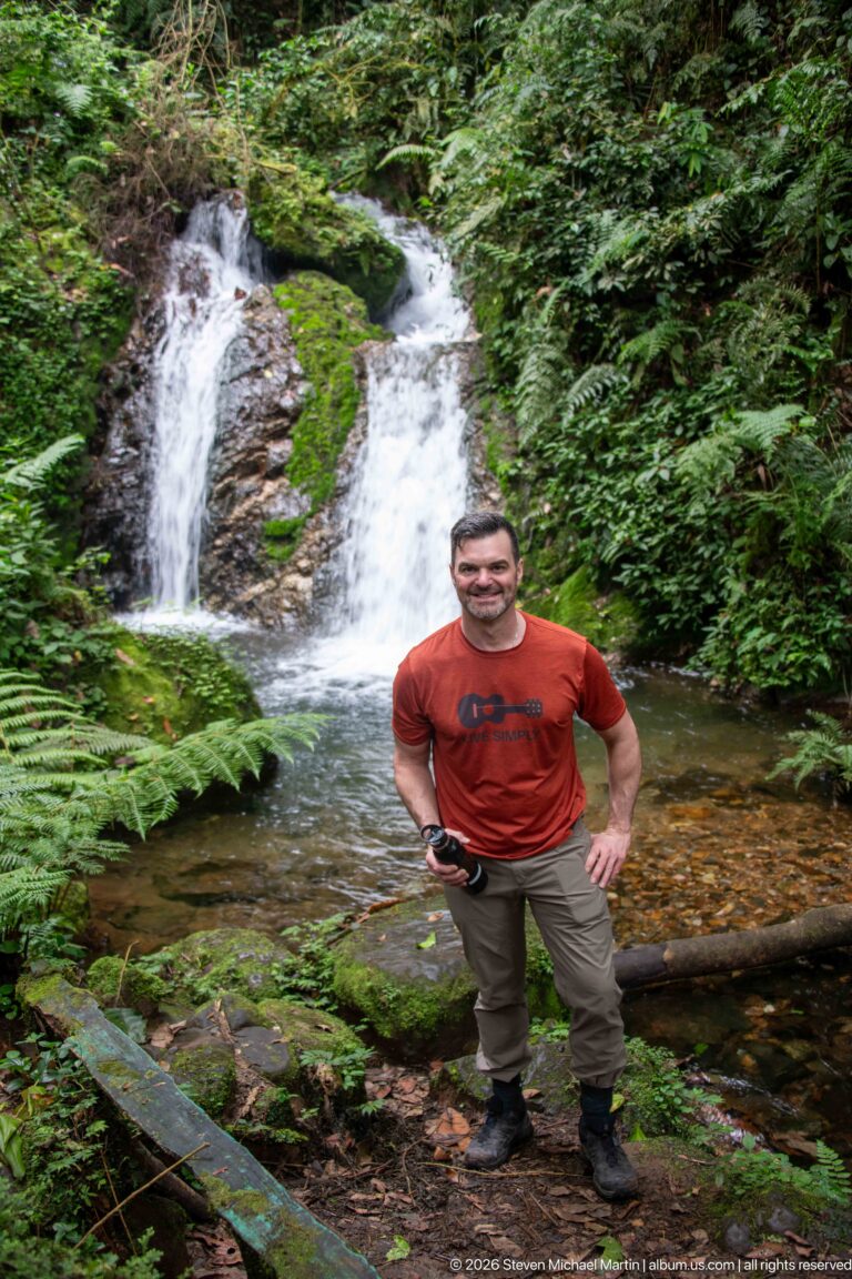 Man in red shirt standing beside a small twin waterfall and forest pool.