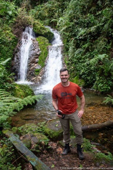 Man in red shirt standing beside a small twin waterfall and forest pool.