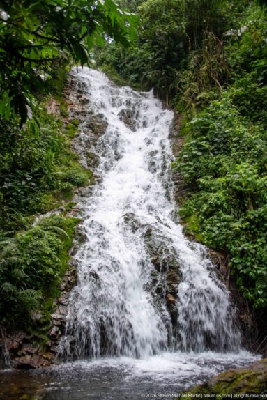 Broad white waterfall descending over dark rock into a small pool amid dense forest.