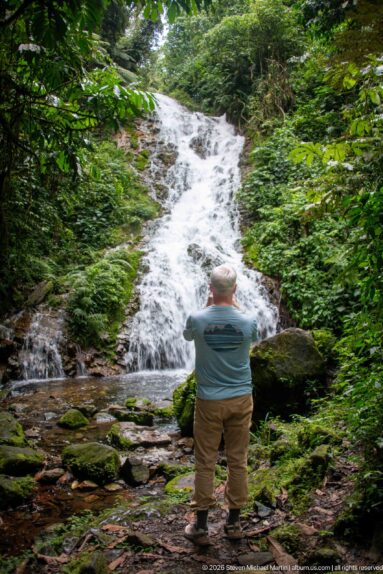 Visitor seen from behind looking toward a tall forest cascade from the rocky streambed.