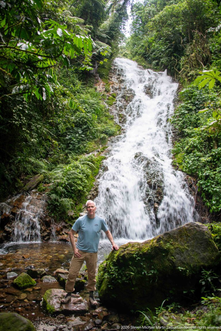 Man standing on rocks below a broad sloping waterfall surrounded by dense green forest.