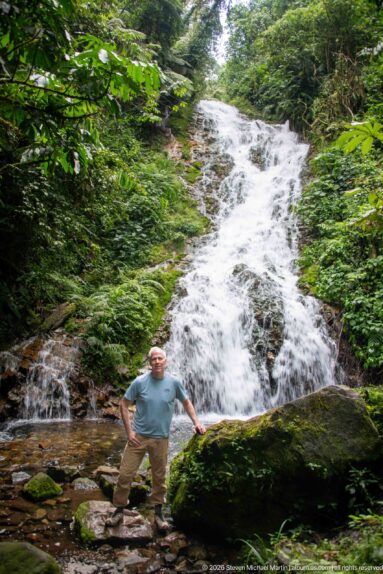 Man standing on rocks below a broad sloping waterfall surrounded by dense green forest.