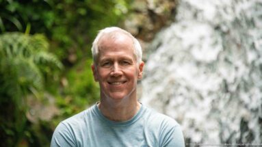 Smiling man in blue shirt framed against a softly focused forest waterfall.