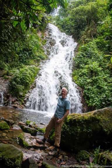 Visitor leans on a mossy fallen trunk with broad Munyaga Falls descending through dense forest behind him.