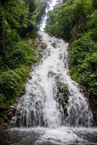 Broad Munyaga Falls descends over a sloping rock face between dense green forest into a dark pool.