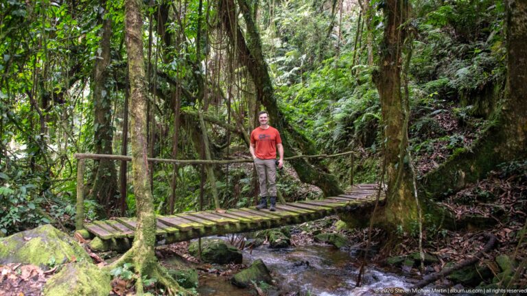 Visitor stands on a narrow wooden footbridge over a rocky stream amid vines and dense forest.