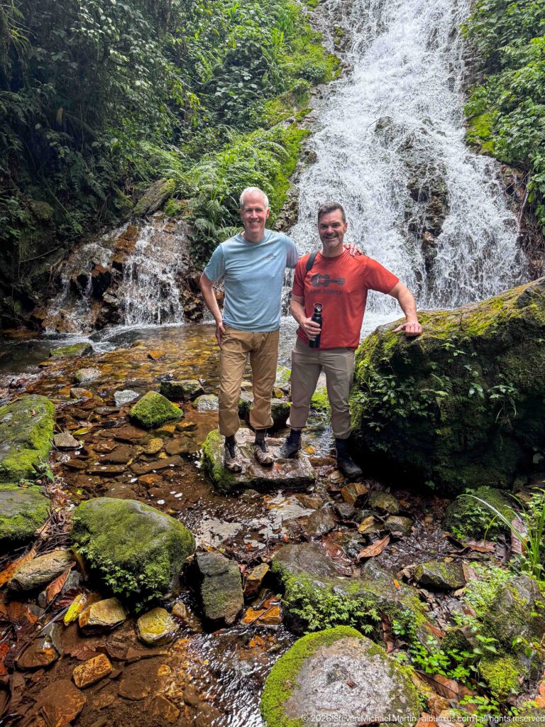 Two visitors stand on rocks in front of Munyaga Falls amid mossy boulders and dense forest.