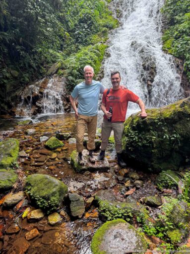 Two visitors stand on rocks in front of Munyaga Falls amid mossy boulders and dense forest.