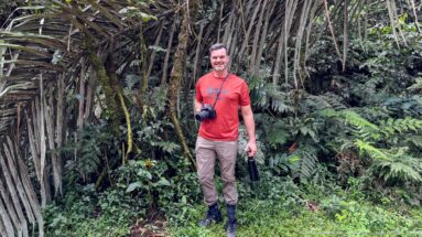 Man in red shirt standing on a forest trail beside dense cane-like stems and ferns.