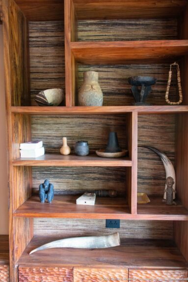 Large wooden shelving unit with small vessels, carved objects and horn-like forms arranged above woven lower storage.