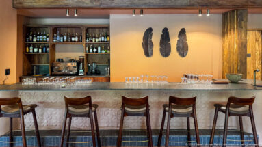 Five bar stools face a pale counter with rows of glasses, bottle shelves, espresso machine and three dark wall sculptures.
