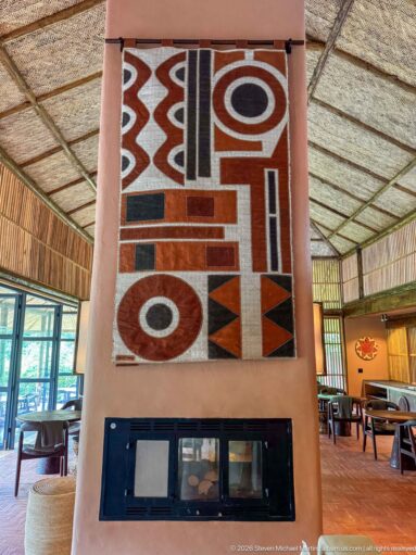 Bold geometric textile in red, black, cream and brown mounted above a dark glazed lodge fireplace.