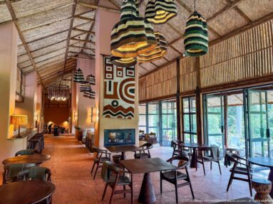 Long lodge interior with dining tables, striped pendant lights, central fireplace and geometric wall textile.