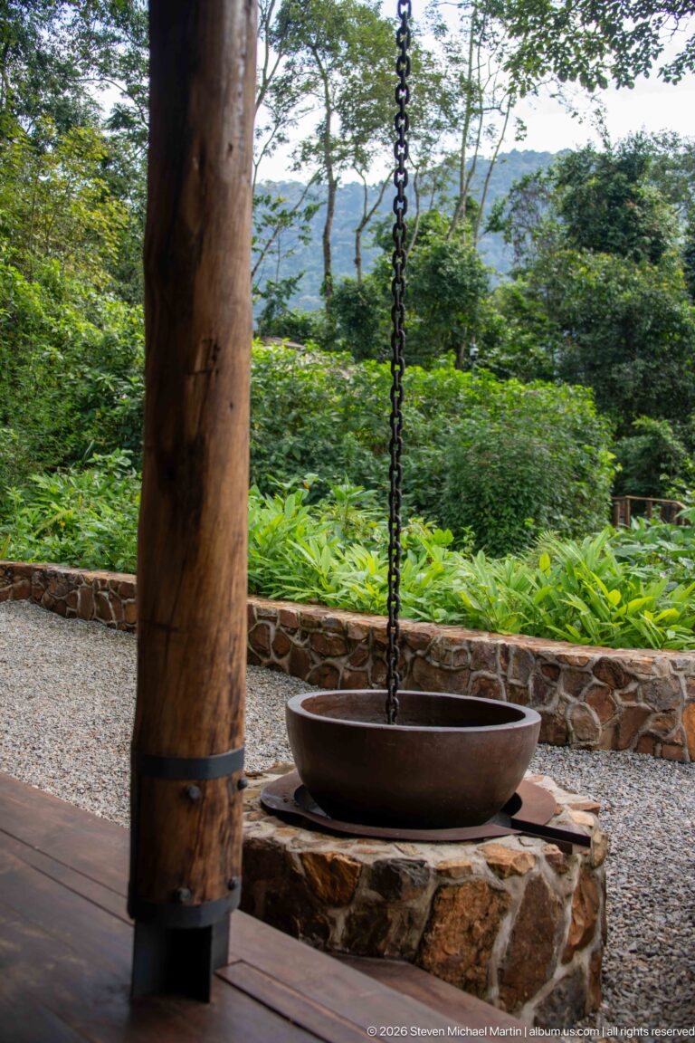 Shallow metal basin suspended by chains beside a large timber veranda post, with garden and wooded hills beyond.