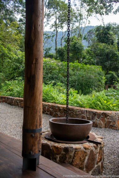 Shallow metal basin suspended by chains beside a large timber veranda post, with garden and wooded hills beyond.