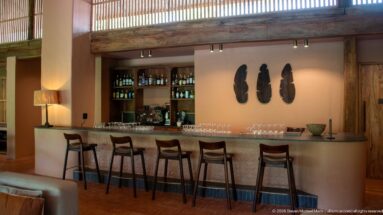 Long lodge bar with five stools, bottle shelves, glassware and three dark sculptural forms mounted on the wall.