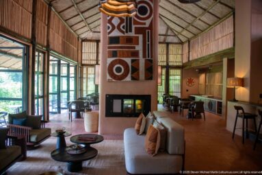 Spacious lodge lounge with sofas, round tables, central fireplace and geometric textile beneath a high woven roof.