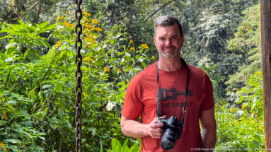 Man in a red shirt holding a camera while standing among dense green vegetation at the lodge.