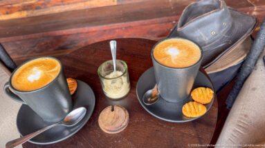 Two gray mugs of foamed coffee with biscuits, spoons and sugar on a wooden table beside a dark brimmed hat.