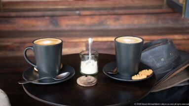 Two dark mugs of foamed coffee with saucers, spoons and a sugar dispenser on a round lodge table.