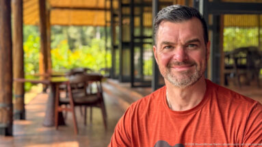 Man in a red shirt facing the camera inside an open lodge interior with tables and greenery softly blurred behind.