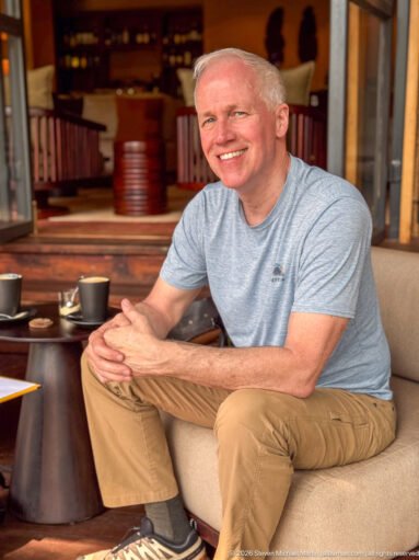 Smiling man seated in a lodge lounge beside a coffee table, with timber shelving and warm interior furnishings behind him.
