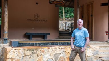Man standing beside the Gorilla Forest Lodge wall sign, dark bench, stone terrace and timber entrance doors.