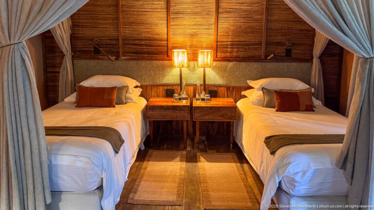 Two neatly made twin beds beneath parted mosquito nets, with matching bedside tables, lamps and a woven floor runner.