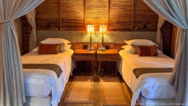 Two neatly made twin beds beneath parted mosquito nets, with matching bedside tables, lamps and a woven floor runner.