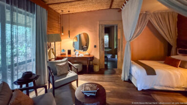 Guest suite with net-draped bed, armchair, desk and round mirror, with an open doorway leading to the bathroom.