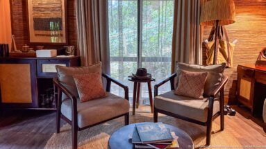 Two cushioned armchairs beside a tall forest-facing window, with a small table, books and timber furnishings.