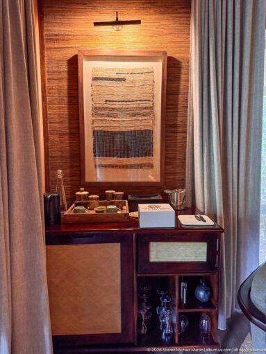 Wooden guest-room refreshment cabinet with cups and glassware beneath framed woven textile artwork and a picture light.