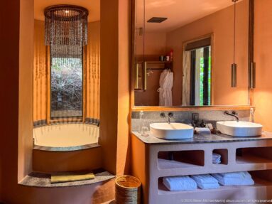 Ochre bathroom with recessed soaking tub, beaded light, double vessel-sink vanity and forest-facing windows.