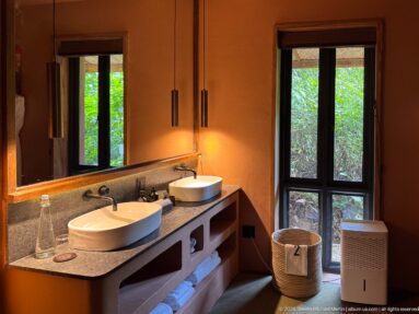 Double vanity with two white vessel sinks, large mirror, folded towels and a glass door opening to forest greenery.