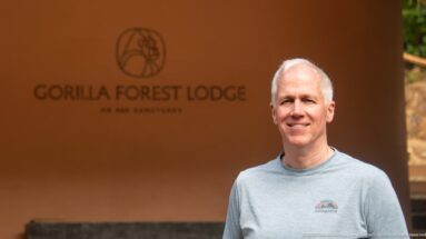 Smiling man in a gray shirt standing before the Gorilla Forest Lodge name and gorilla emblem on an ochre wall.