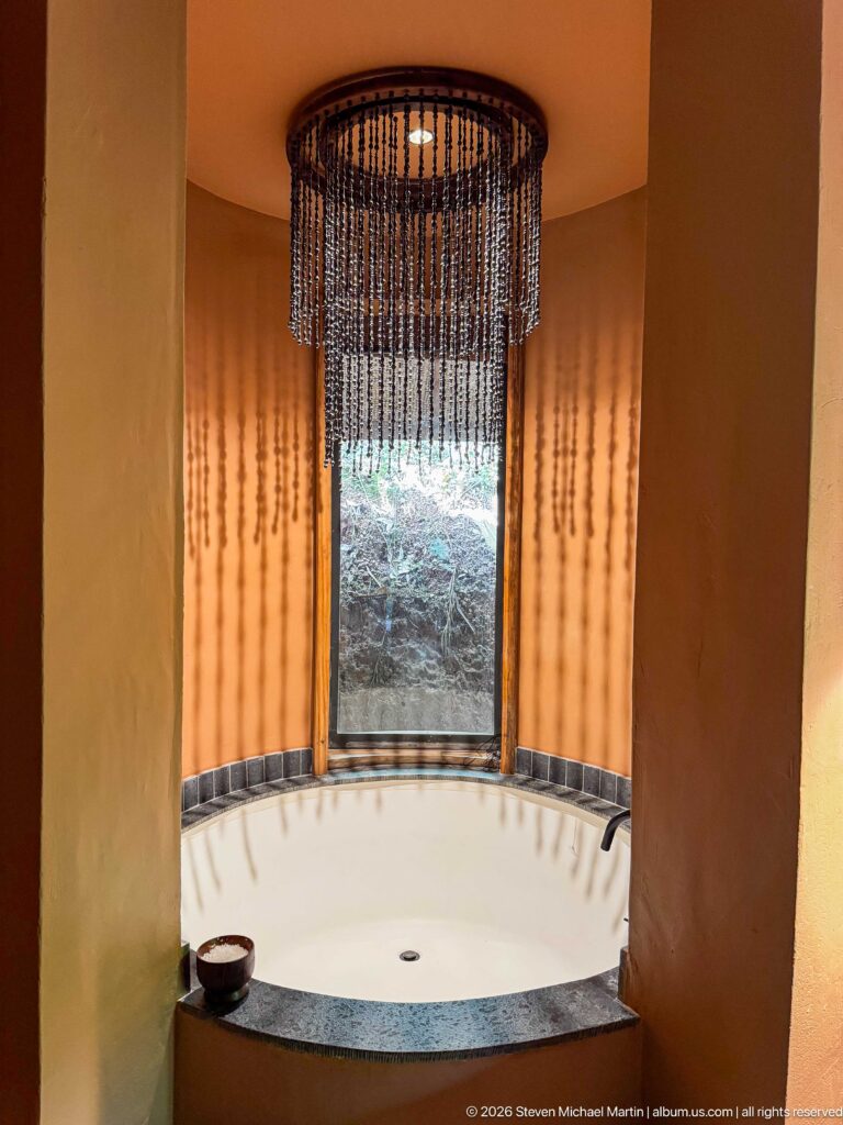 Recessed soaking tub in an ochre alcove beneath a beaded pendant light, with a narrow window facing vegetation.